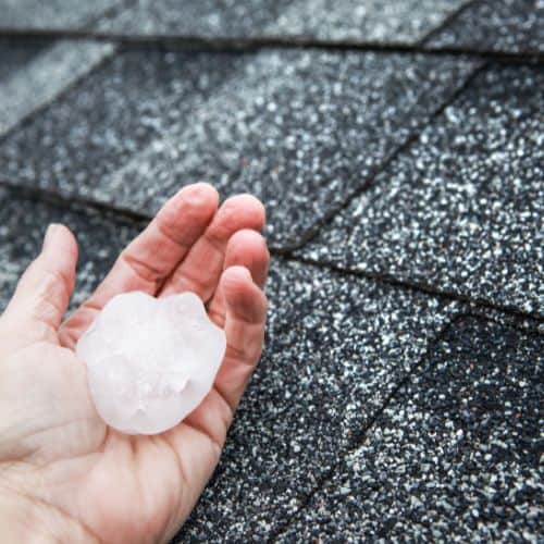 a hand holding a large piece of hail in front of a roof that may have storm damage